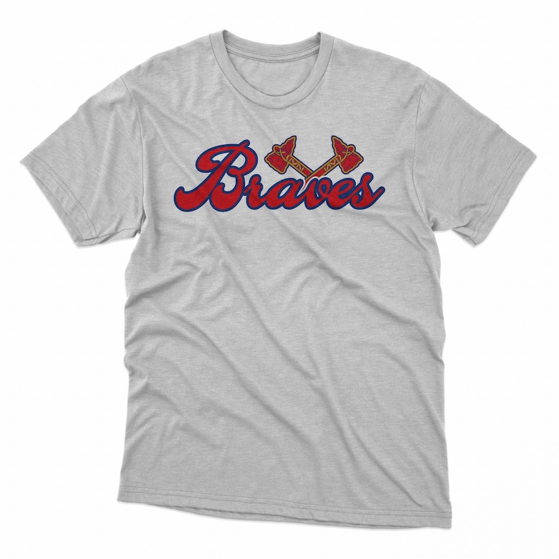 Puede incluir: Camiseta gris claro con la palabra "Braves" en rojo con un contorno azul. Encima de la palabra hay dos tomahawks cruzados en rojo y dorado.