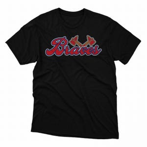 Puede incluir: Camiseta negra con la palabra "Braves" en escritura roja con un contorno azul. Encima de la palabra hay dos tomahawks cruzados en rojo y dorado. La camiseta est&aacute; hecha de un material suave.