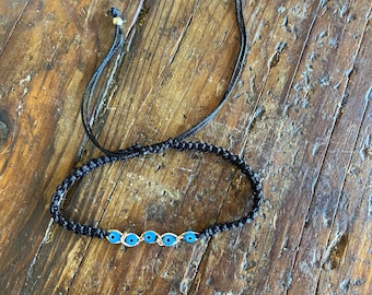 May include: A black string bracelet with five blue evil eye charms. The bracelet has adjustable string ties.