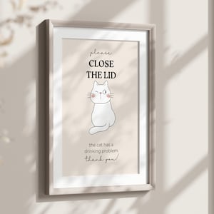 May include: Framed art print with a light beige background. The print features a winking white cat illustration and the text "please CLOSE THE LID" above it. Below the cat, the text reads "the cat has a drinking problem! thank you!"