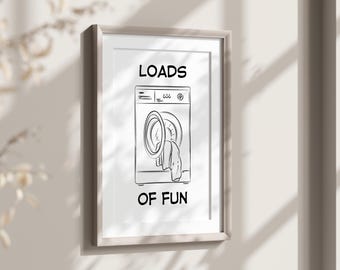 May include: A framed black and white print featuring a line drawing of a washing machine with the words "LOADS OF FUN" in bold letters. The artwork is mounted in a light-colored wooden frame and hangs on a neutral-toned wall.