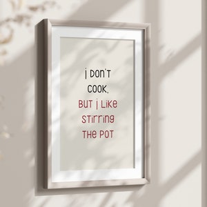May include: Framed art print with the text "I don't cook, but I like stirring the pot" in black and red lettering. The print is in a light-colored frame and hangs on a neutral-colored wall.
