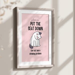 Peut inclure: Impression encadrée avec un fond rose et une illustration de chat de dessin animé. Le texte se lit "please PUT THE SEAT DOWN the cat has a drinking problem" en noir. Le cadre est gris clair.