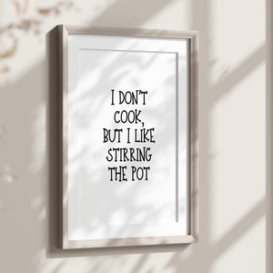 May include: Framed print with the text "I don't cook, but I like stirring the pot" in black lettering. The print is in a light-colored frame and hangs on a neutral-colored wall. The overall aesthetic is minimalist and modern.
