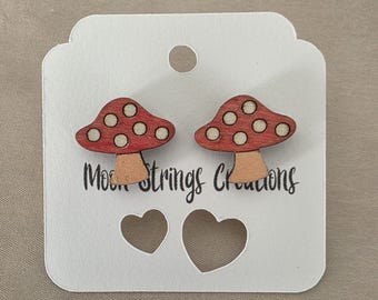 May include: A pair of wooden mushroom earrings with red caps and white spots. The earrings are displayed on a white card with the text "Moon Strings Creations" and heart-shaped cutouts.