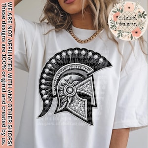May include: White t-shirt featuring a detailed black and silver graphic of a Spartan helmet. The design includes sparkling embellishments and intricate detailing. Text on the shirt reads "WE ARE NOT AFFILIATED WITH ANY OTHER SHOPS! These designs are 100% original and created by us."