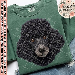 May include: A green Comfort Colors sweatshirt featuring a detailed illustration of a black poodle's face, overlaid with a grid pattern. The poodle has brown eyes and a textured coat. The text "original designer" is in a floral circle.