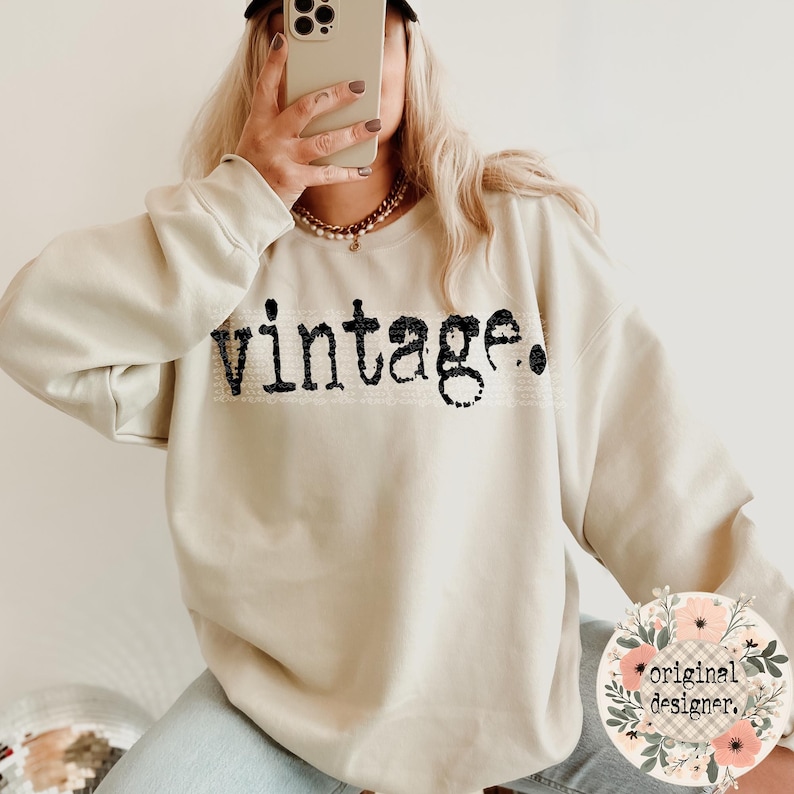A person in a light beige sweatshirt with "vintage." in black distressed text across the chest, holding a phone to their face, with a floral "original designer." logo in the corner.