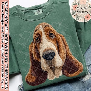 Puede incluir: Una sudadera verde con una imagen detallada de la cara de un Basset Hound. El pelaje del perro es marrón, beige y blanco, con orejas largas y caídas. La sudadera tiene una etiqueta Comfort Colors y el texto "original designer."