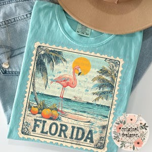 May include: A light blue t-shirt featuring a graphic print of a pink flamingo standing on a surfboard near palm trees and oranges. The design is framed like a postage stamp with the text FLORIDA at the bottom. The shirt has a crew neck collar.