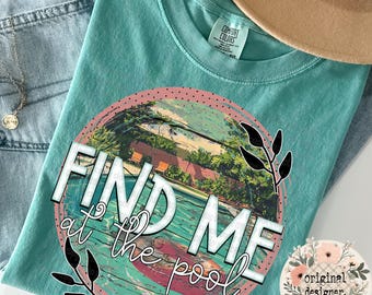 May include: Teal Comfort Colors t-shirt with a graphic that says "FIND ME at the pool." The graphic features a pool scene with a person on a float. The shirt is laid on top of blue jeans and a tan hat.