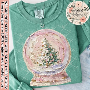 May include: A teal Comfort Colors t-shirt featuring a large, textured painting of a snow globe with a Christmas tree inside. The snow globe is painted in shades of pink, white, and gold. The shirt also has a silver necklace and the text "original designer."