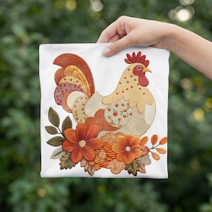 Op de afbeelding: Een wit t-shirt met een geborduurd ontwerp van een haan met beige, oranje en rode veren. De haan is omringd door oranje en rode bloemen met groene bladeren. Het borduurwerk gebruikt verschillende texturen en patronen voor de veren.