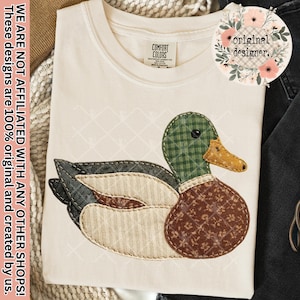 May include: A cream-colored t-shirt featuring a large mallard duck applique. The duck is constructed from various fabric patches including green gingham, brown floral print, and cream ribbed textures, all outlined with visible dark stitching.