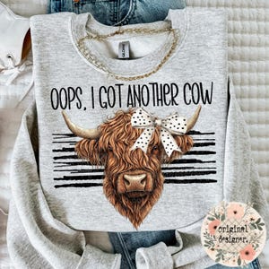 Könnte beinhalten: Ein graues Sweatshirt mit dem Text "Oops, I got another cow" darauf gedruckt. Ein braunes und weißes Highland-Rind mit einer schwarz-weißen Polka-Dot-Schleife ist auf dem Sweatshirt abgebildet.