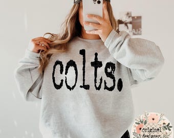 May include: A gray sweatshirt with the word "colts" printed in black on the front. The sweatshirt is being worn by a person with long brown hair.