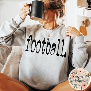 May include: A light gray crewneck sweatshirt featuring the word football printed in a distressed black font across the chest. The garment has long sleeves and a ribbed neckline, cuffs, and hem, providing a casual and comfortable fit for daily wear.