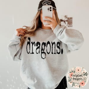 May include: A gray sweatshirt with the text "Dragons." printed in black. The sweatshirt is being worn by a person with a black baseball cap.