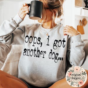 May include: A light gray crewneck sweatshirt featuring the black text "oops, i got another dog..." printed across the chest. The garment has long sleeves and a ribbed neckline, cuffs, and hem, providing a casual look for everyday wear.