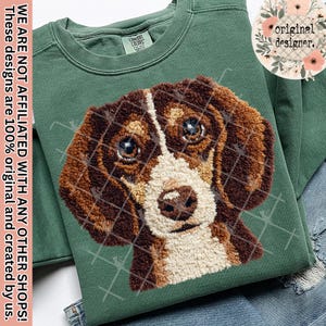 Può includere: Una felpa verde con un'immagine dettagliata e strutturata del muso di un cane. Il cane ha il pelo marrone e bianco, con un motivo a griglia sovrapposto al disegno. Il testo "original designer" è in un cerchio floreale.