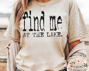 Peut inclure: T-shirt beige avec l'inscription "find me at the lake" en noir vieilli. Le t-shirt a des manches courtes et une coupe décontractée. Un motif floral avec les mots "original designer" est visible en bas à droite.