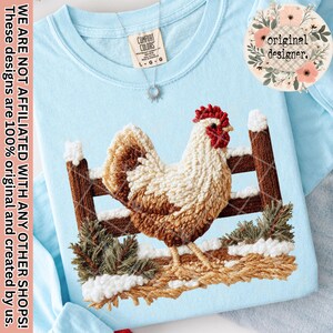 Könnte beinhalten: Hellblaues Comfort Colors Sweatshirt mit einem gestickten Hahn-Design. Der Hahn ist braun, weiß und rot und steht vor einem Holzzaun mit Schnee und Grün. Der Text "original designer" ist sichtbar.