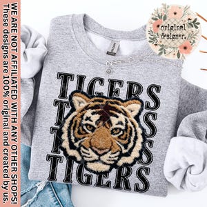 May include: A heather gray sweatshirt with the word "TIGERS" in black block letters and a detailed tiger face graphic. The sweatshirt has a crew neck and long sleeves. A silver necklace with a small pendant is visible.