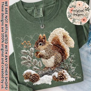 May include: A green long-sleeve shirt featuring an embroidered squirrel design. The squirrel is brown and white, perched on a snowy branch with pinecones. The shirt has the text "original designer."