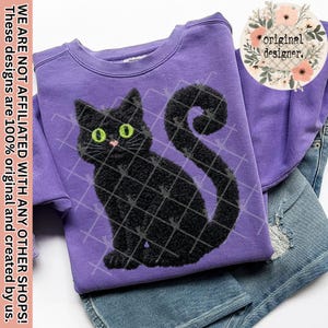 May include: A purple sweatshirt featuring a black cat design with green eyes and a pink nose. The cat is outlined with a grid pattern. The text "original designer" is visible. The sweatshirt is paired with blue denim shorts.