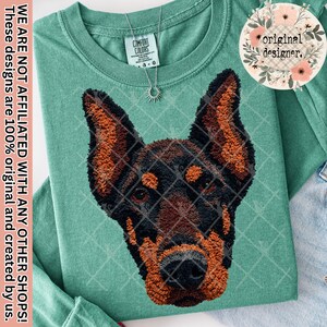 May include: A teal long-sleeved shirt with an embroidered Doberman Pinscher design. The dog's head is black and brown, with a diamond pattern. The shirt has a Comfort Colors label and a sun pendant. The text on the shirt says "original designer."