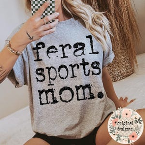 May include: A heather grey short-sleeved t-shirt featuring the black text "feral sports mom." printed in a distressed, textured font across the chest. The shirt has a crew neckline and a relaxed fit, showcasing a casual style with bold lettering.