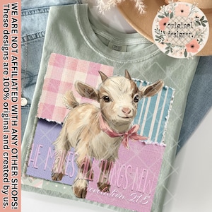 May include: A light green t-shirt featuring a watercolor illustration of a baby goat wearing a pink ribbon. The design includes patchwork squares with text that reads "HE MAKES ALL THINGS NEW Revelation 21:5".