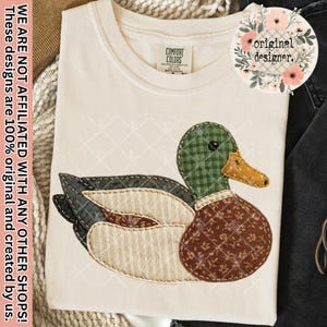 May include: Off-white t-shirt with an appliqued duck design. The duck has a green plaid head, brown patterned body, and a yellow beak. The shirt has the text "COMFORT COLORS" and "original designer."