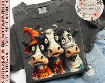 May include: A charcoal gray crewneck sweatshirt featuring a graphic of three cows with wide eyes. One cow wears an orange witch hat, one has a red scarf, and one is wrapped in white bandages. Small black bats are printed above the cows.