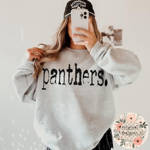 A person wears a gray crewneck sweatshirt featuring the distressed black word "panthers." on the front, with a floral "original designer" logo in the bottom right corner.