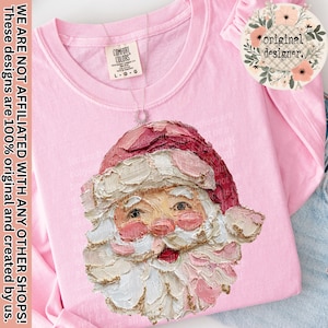 May include: A pale pink long-sleeved shirt with a detailed Santa Claus face design. The Santa Claus face is painted with pink, white, and gold tones. The shirt has a Comfort Colors tag and a necklace. The text "WE ARE NOT AFFILIATED WITH ANY OTHER SHOPS! These designs are 100% original and created by us." is printed on the left side.