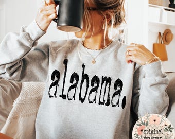 May include: A light gray long-sleeve crewneck sweatshirt featuring the word alabama. printed in a distressed black font across the chest. The garment has a ribbed neckline, cuffs, and hem, and is worn with a gold chain necklace and rings.