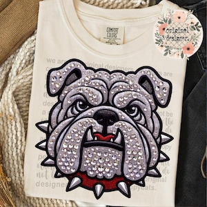 May include: A cream-colored t-shirt featuring a large, central graphic of a bulldog head embellished with clear rhinestones. The bulldog has a red spiked collar and a red mouth. The shirt includes a Comfort Colors label and faint text in the background.