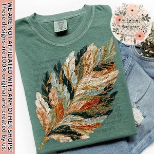May include: Sage green t-shirt featuring a large, textured leaf design with gold glitter accents. The leaf incorporates shades of white, orange, and green. The shirt has the text "Comfort Colors" on the tag and the text "original designer."