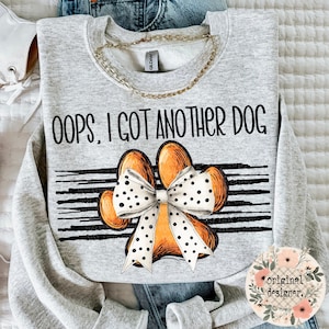A light gray sweatshirt with the text "Oops, I Got Another Dog" above a large orange paw print with a white polka-dotted bow, on a striped background.