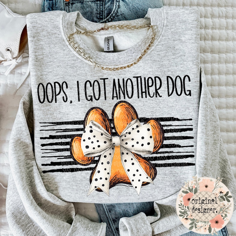 A light gray sweatshirt with the text "Oops, I Got Another Dog" above a large orange paw print with a white polka-dotted bow, on a striped background.