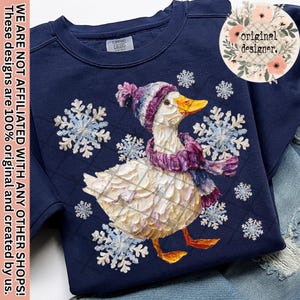 May include: Navy blue sweatshirt featuring a whimsical illustration of a duck wearing a winter hat and scarf, surrounded by snowflakes. The duck has a yellow beak and is set against a quilted background. The text "original designer" is in a floral circle.