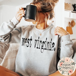 A person wearing a gray sweatshirt with the distressed black text west virginia across the chest, holding a black mug.