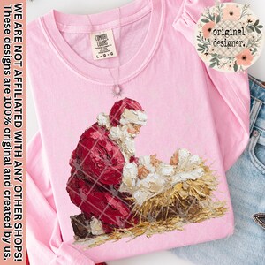 May include: A light pink long-sleeve shirt featuring a painting of Santa Claus kneeling beside baby Jesus in a manger. The shirt has a Comfort Colors tag and the text "original designer."