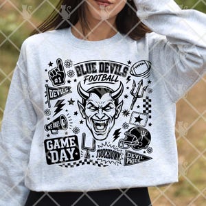 May include: A light gray long-sleeve sweatshirt featuring a black graphic print of a devil face surrounded by football-themed icons including a #1 foam finger, a football, a megaphone, a goal post, and text reading Blue Devils Football.