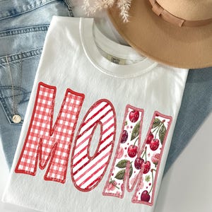 May include: White t-shirt with the word "MOM" in red. The "M" is red gingham, the "O" is red and white striped, and the "M" is decorated with cherries and floral accents. The shirt is on top of blue jeans.