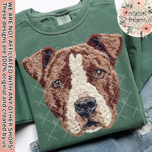 May include: Green sweatshirt with an embroidered dog face in brown, white, and tan. The dog's eyes are brown, and the design is centered on the shirt. The text "original designer" is in a floral circle. The shirt is folded on a pair of jeans.