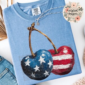 May include: A blue and red cherry design on a light blue t-shirt. The cherries are painted in the style of the American flag.