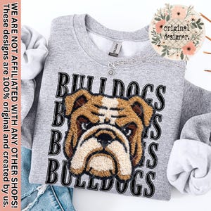 Peut inclure: Un sweat-shirt gris avec un motif de bouledogue et le mot "BULLDOGS" superposé. Le motif est marron, beige et noir. Le sweat-shirt a un col rond et des manches longues. Un collier argenté avec un pendentif soleil est visible.