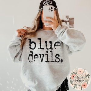 May include: A gray sweatshirt with the text "blue devils" printed in black. The sweatshirt is being worn by a person with a black baseball cap.